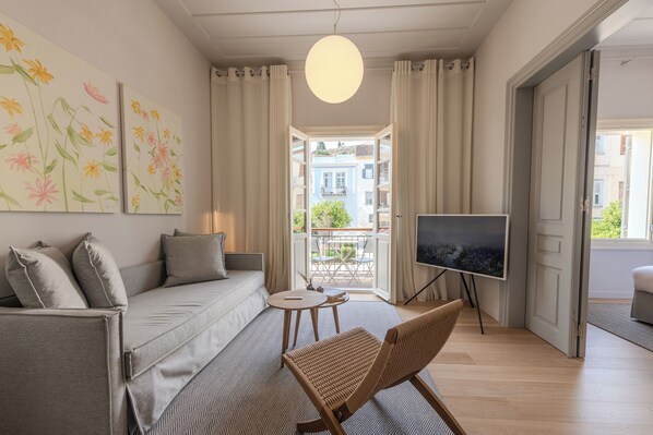A modern living room with a sofa, a TV on a stand, a wicker chair, and a balcony with a view of buildings. - Amalie Suites (Nafplio)