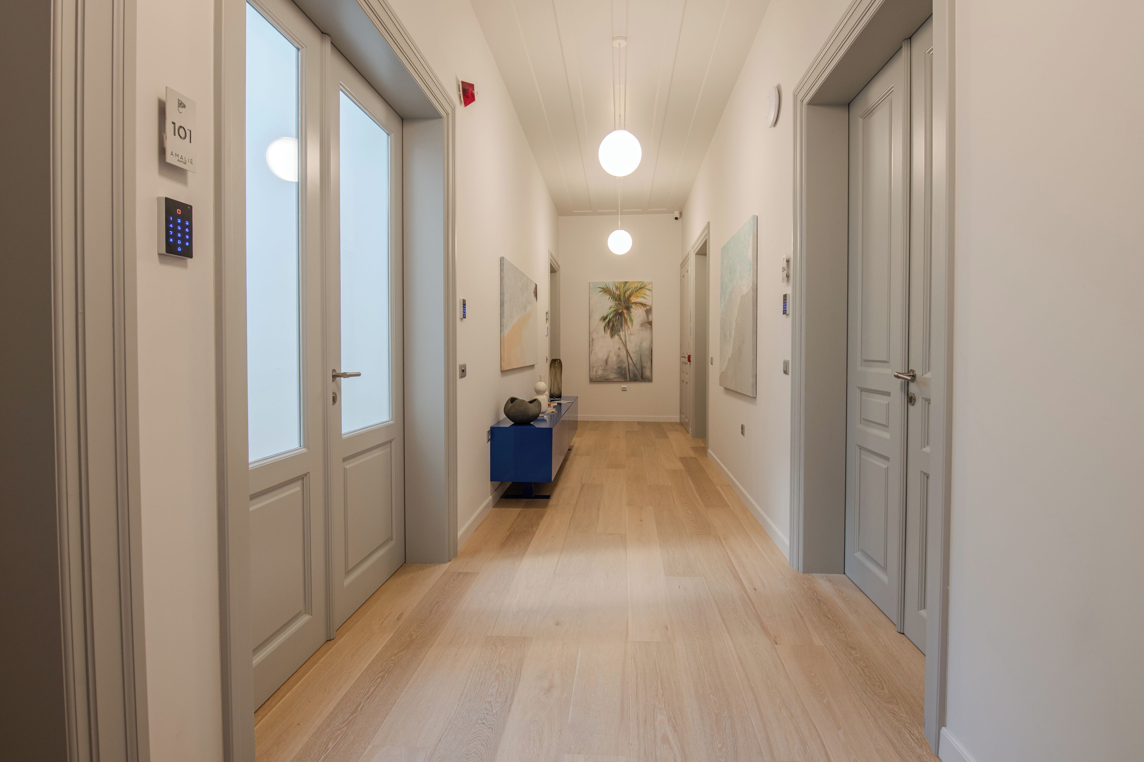 Un pasillo con piso de madera, paredes blancas y un banco azul. Hay puertas a ambos lados y una lámpara cuelga del techo.