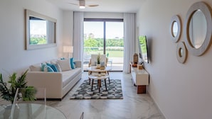 Un salon moderne avec un canapé, une table basse et une table à manger. Il y a une grande fenêtre avec des rideaux, un ventilateur de plafond et un téléviseur posé sur un meuble.