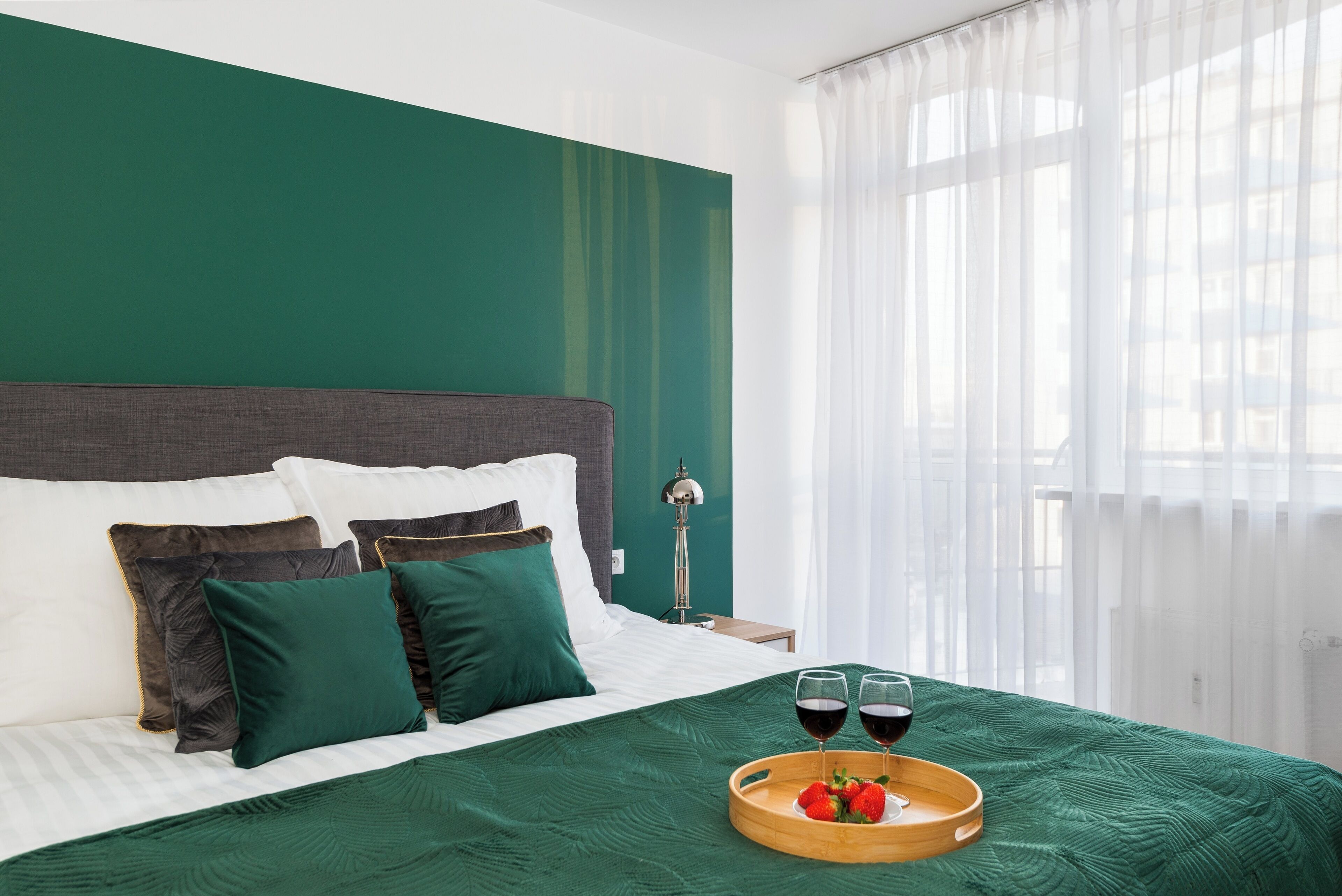 A neatly made bed with green bedding, two glasses of red wine, and a tray with strawberries on a wooden tray.