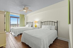 A hotel room with two beds, a ceiling fan, a nightstand with a lamp, and a view of the beach.