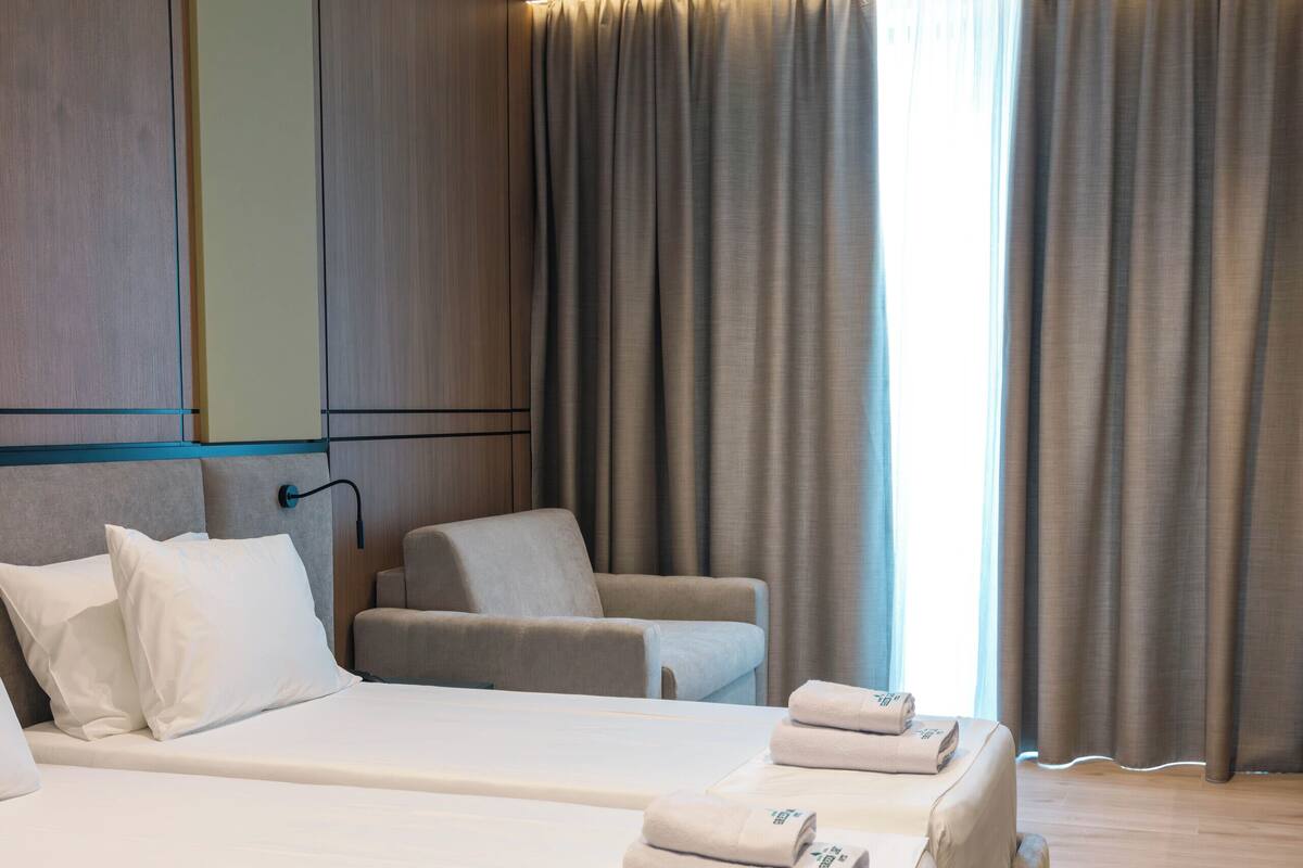 Una habitación de hotel moderna con dos camas, un sofá y un amplio ventanal con cortinas.