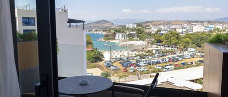 A balcony with a table and chairs, overlooking a coastal town with a beach, parked cars, and buildings.