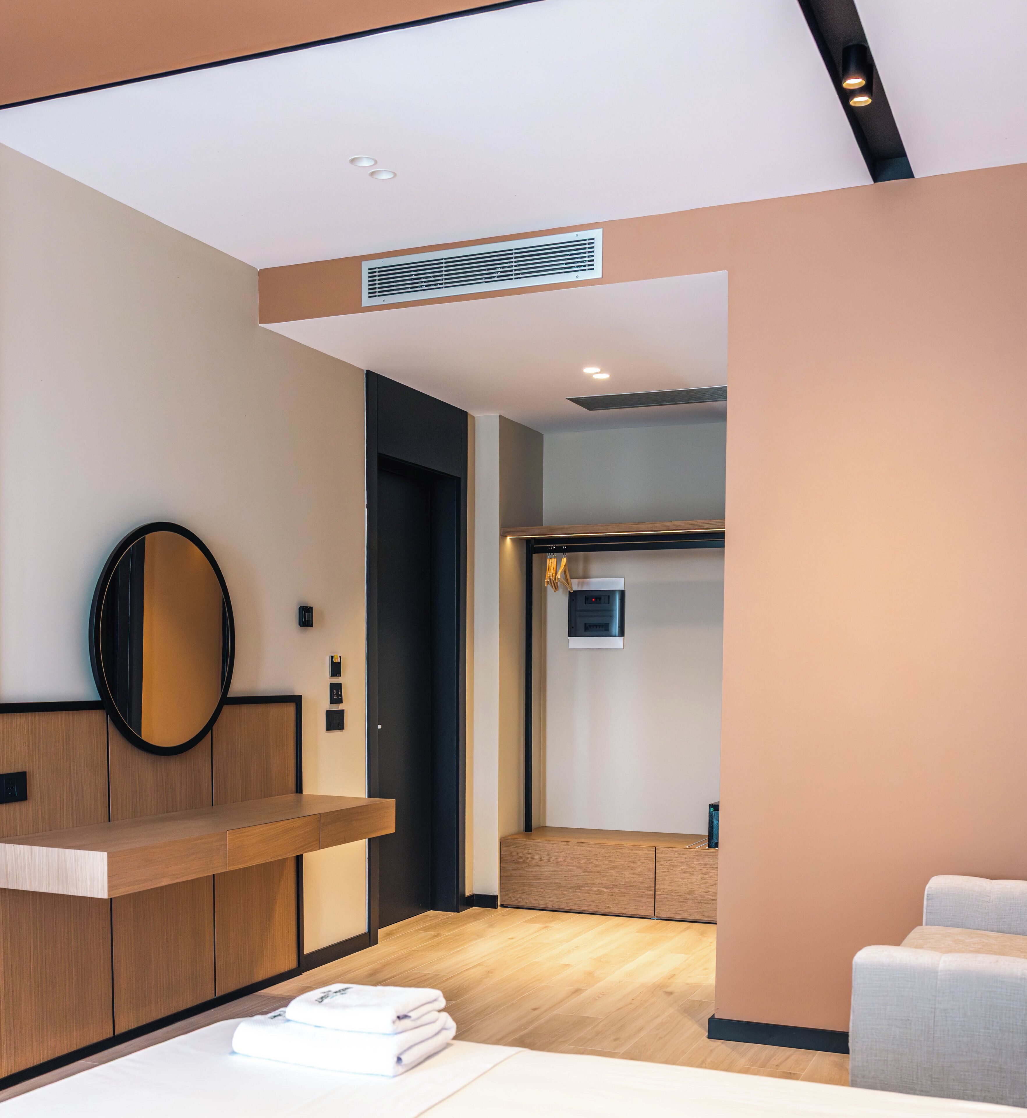 Une chambre d’hôtel moderne avec un comptoir d’accueil en bois, un grand miroir, un lit avec une serviette blanche soigneusement pliée, et une vue sur le couloir.