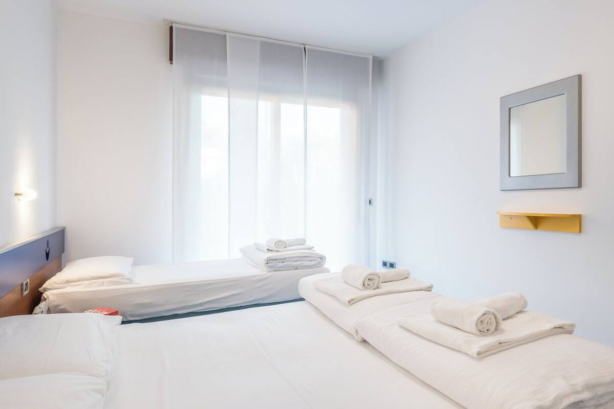 Ein Hotelzimmer mit zwei Betten, einem großen Fenster mit durchsichtigen Vorhängen, einem Nachttisch mit Lampe und einem gerahmten Bild an der Wand.