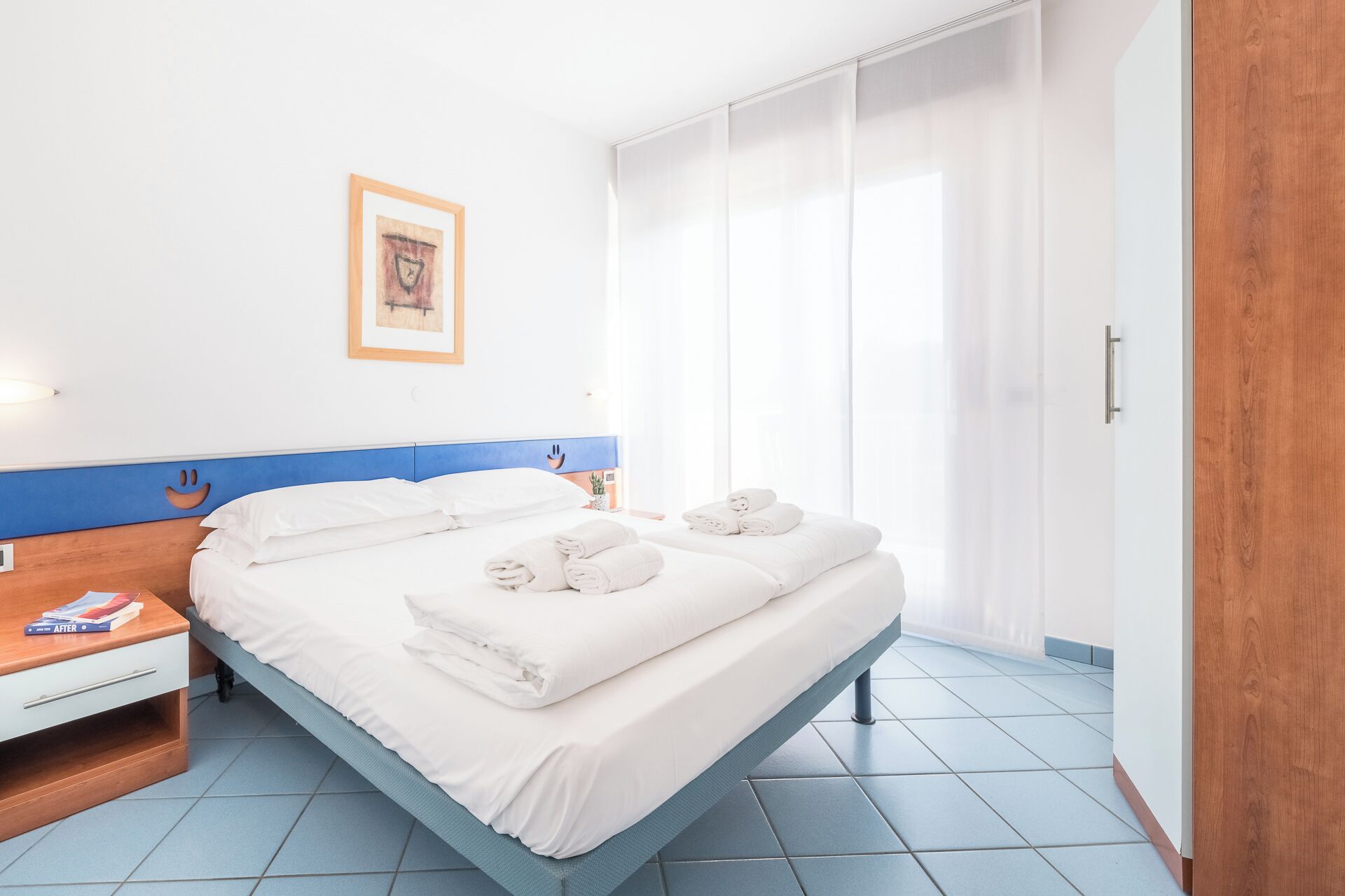 A hotel room with a bed, a bedside table with a book, a wardrobe, and a window with curtains.