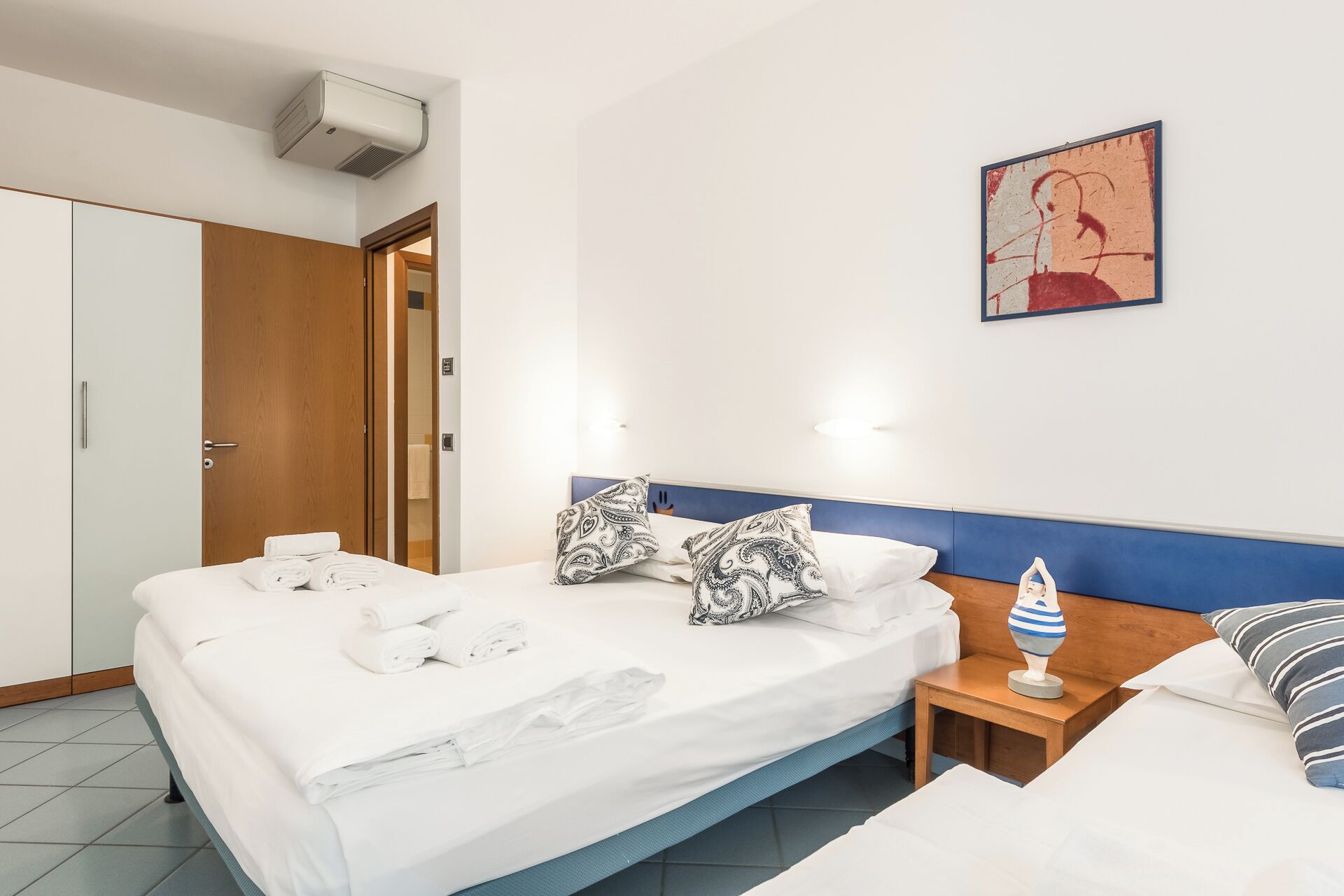 Una habitación de hotel con una cama, una mesita de noche de madera, un aire acondicionado de pared y un cuadro.