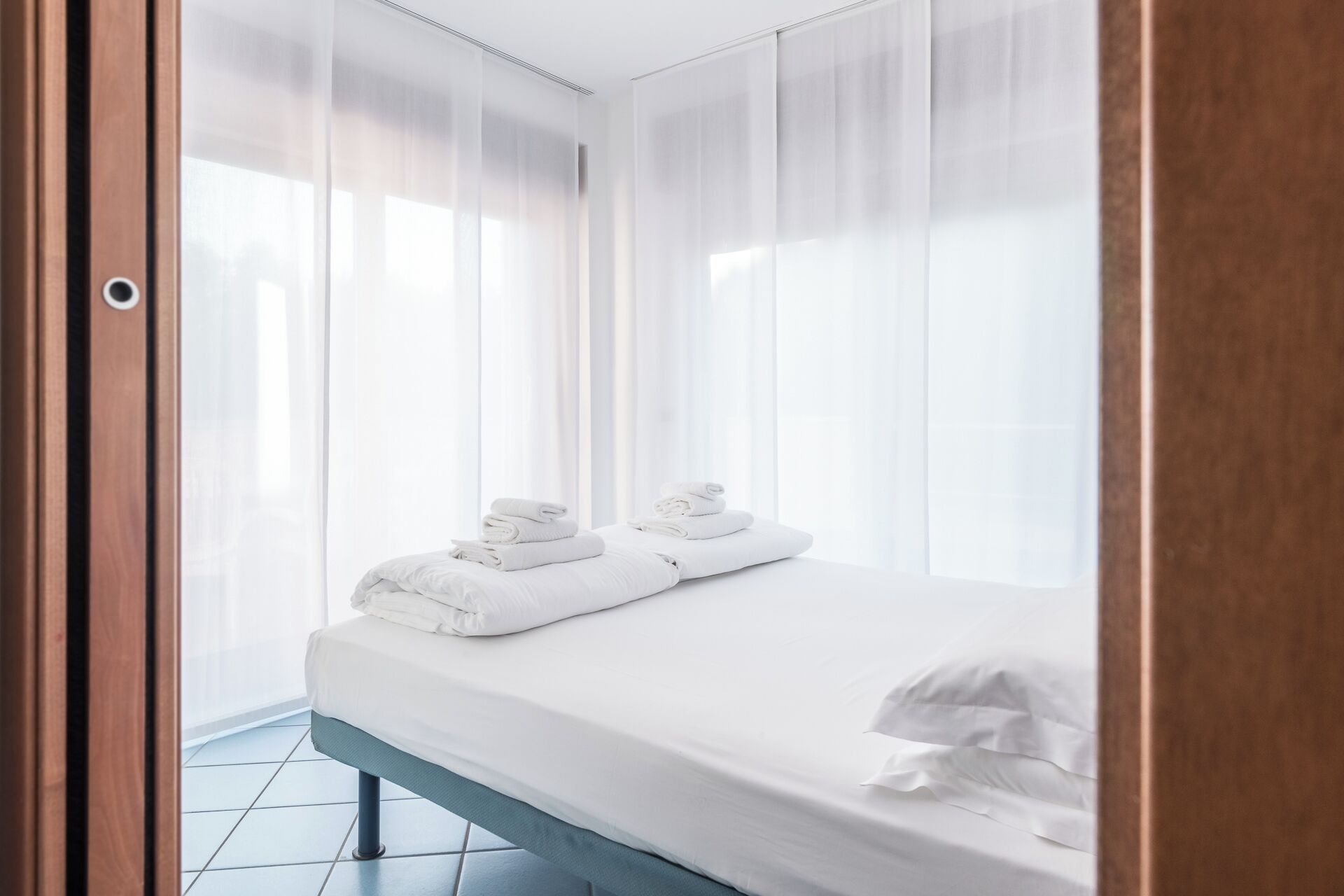 A neatly made bed with white linens and towels, a wooden door frame, and a tiled floor.