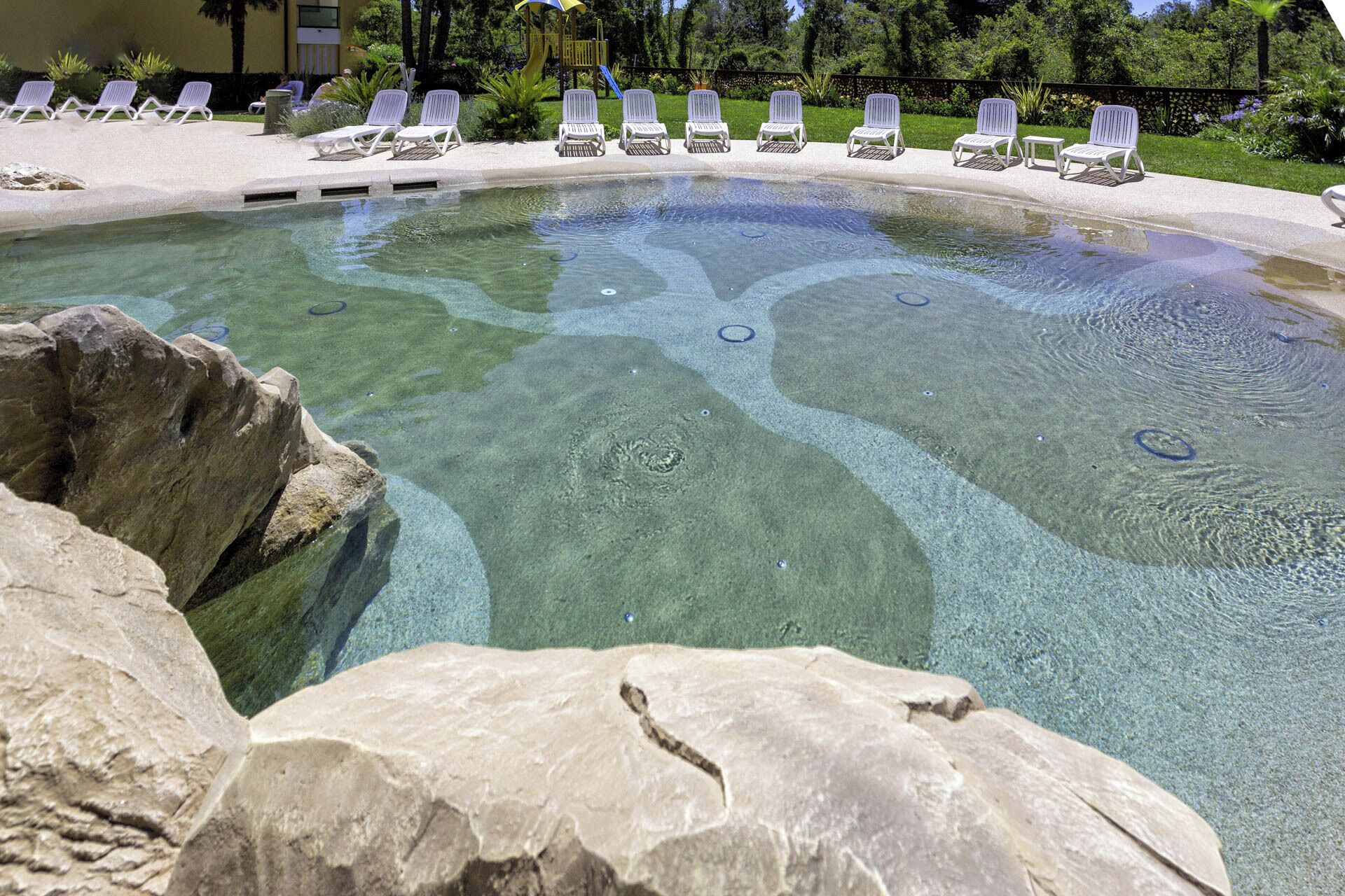 Una piscina limpida con lettini bianchi tutt'intorno, circondata da rocce e vegetazione.