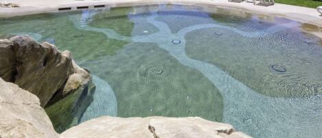 Una piscina limpida con lettini bianchi tutt'intorno, circondata da rocce e vegetazione.