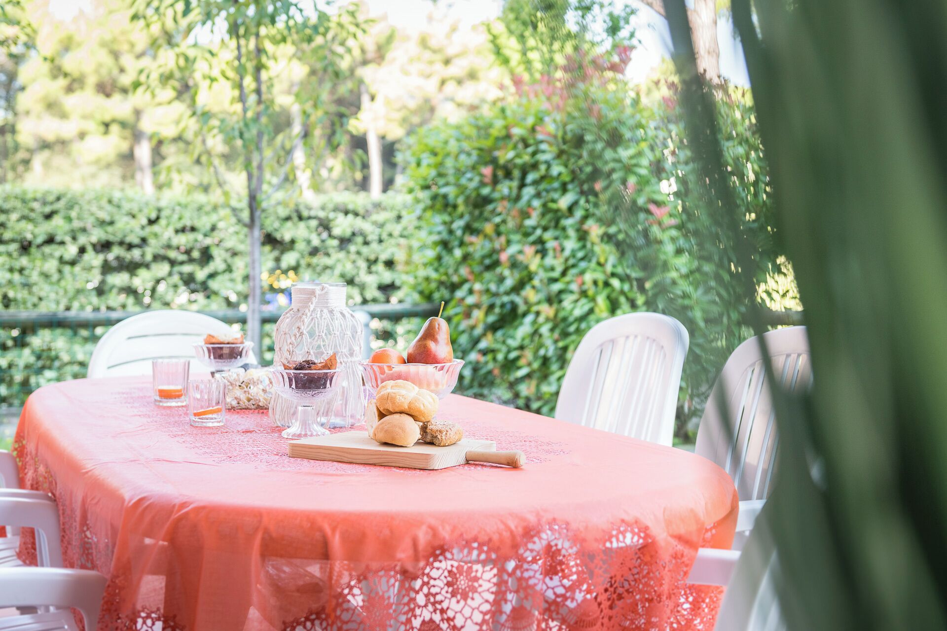 Una mesa de comedor al aire libre con mantel rojo, cristalería, pasteles y una cesta de frutas.