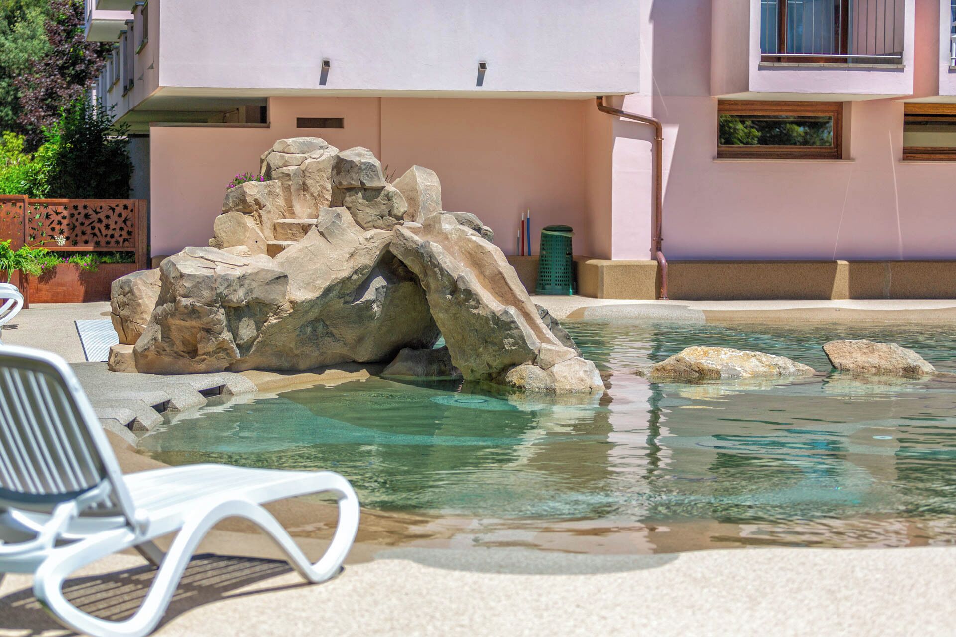 Zona de piscina con una formación rocosa, una silla de descanso blanca y un edificio al fondo.
