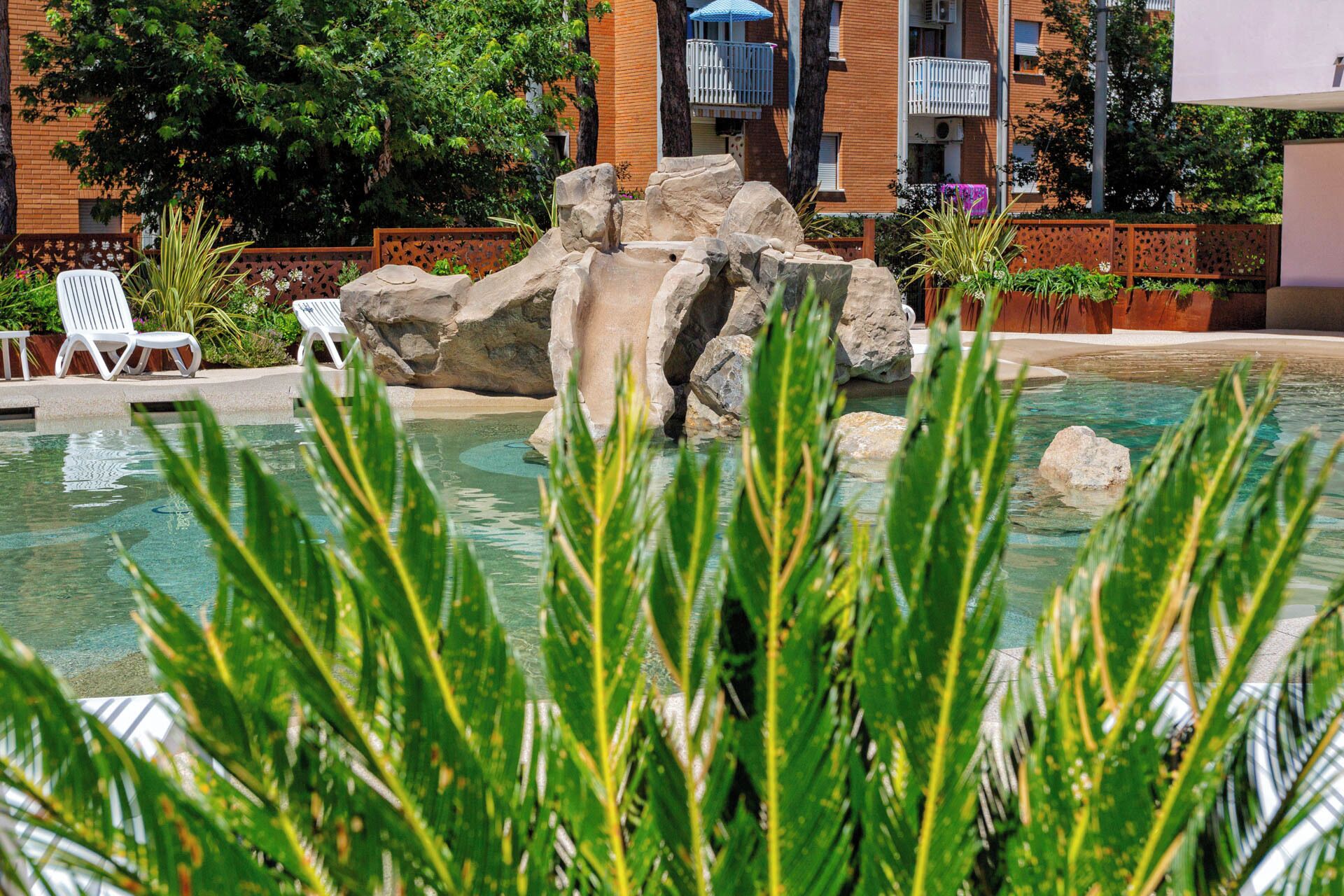 Une piscine avec une cascade artificielle en roche, entourée de verdure et de chaises longues blanches.