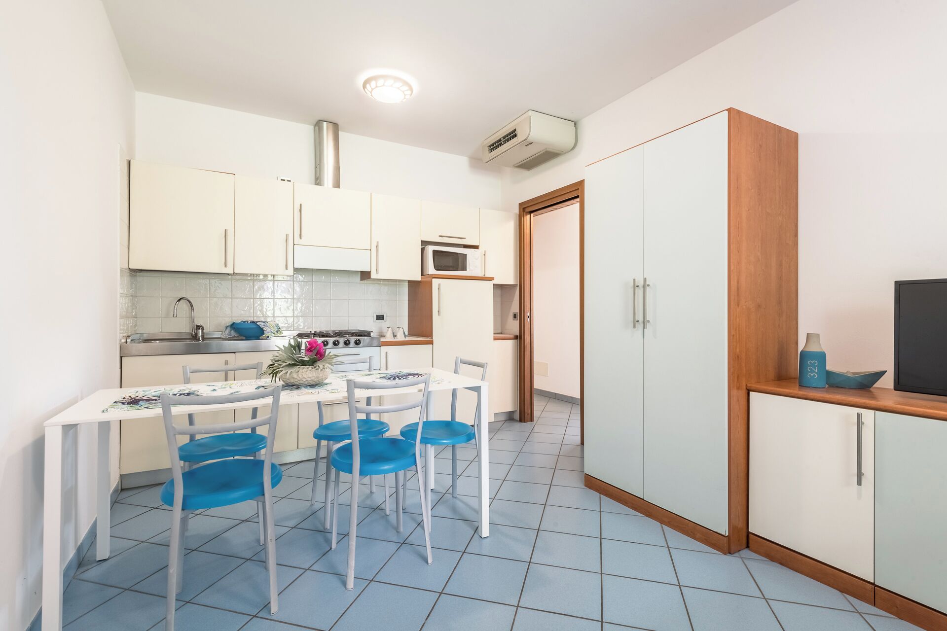 A compact kitchen with white cabinets, a microwave, and a stove. There is a dining table with blue chairs and a small TV on a wooden shelf.