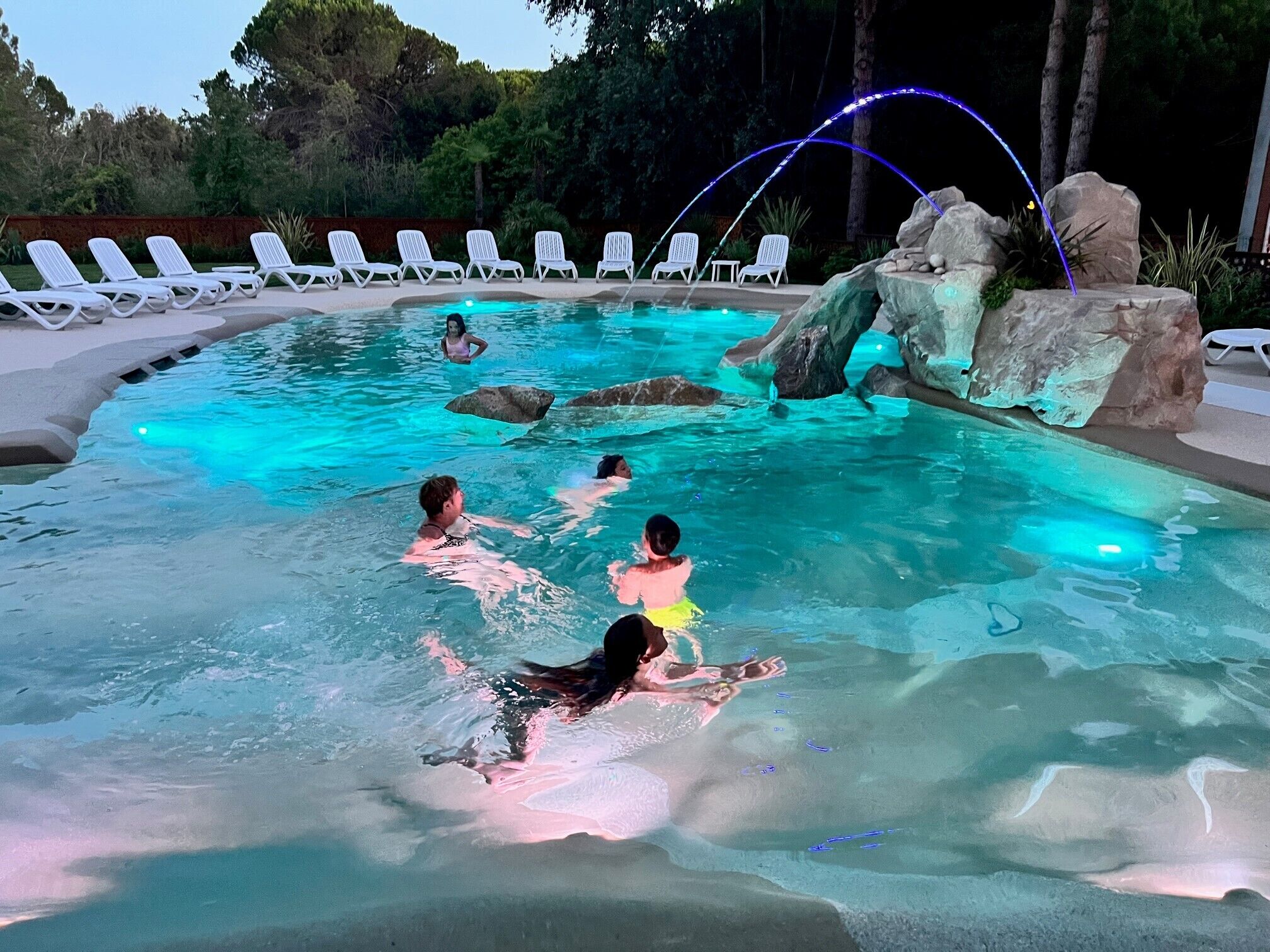 A swimming pool with clear water, surrounded by white lounge chairs and a rock feature.