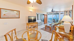 A spacious living area with a television, wicker furniture, and a balcony with a view. - Oceanfront Bliss: Pool, Hot Tub and Private Lanai (Kailua-Kona)