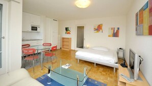 A compact living space with a kitchenette, a dining area with red chairs, a bed, a TV, and abstract wall art. - Spacious studio with balcony (Vineyard 71) (Zürich)