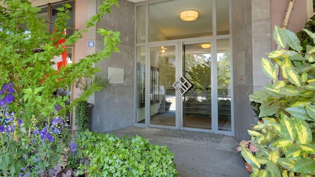 A building entrance with a glass door, a small outdoor seating area, and various plants. - Spacious studio with balcony (Vineyard 71) (Zürich)