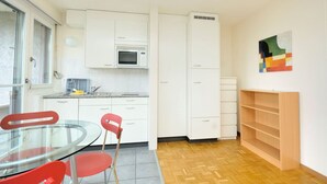 A compact kitchen with white cabinets, a microwave, and a built-in oven. There's a dining area with a glass table and red chairs, and a wooden shelf unit against the wall. - Spacious studio with balcony (Vineyard 71) (Zürich)