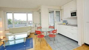 A modern apartment with a glass dining table, red chairs, a white sofa, and a kitchen with white cabinets. - Spacious studio with balcony (Vineyard 71) (Zürich)