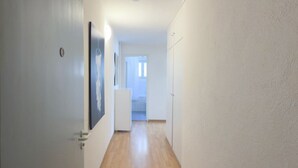 A narrow hallway with wooden flooring and white walls. There are framed pictures on the left wall and a door at the end.