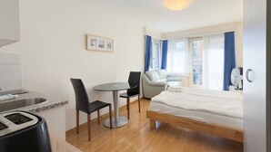 A modern hotel room with a bed, a dining table with chairs, a small kitchen area, and a window with curtains.