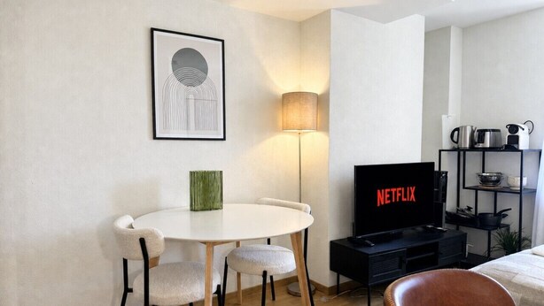 A modern living room with a round dining table, a sofa, a TV displaying 'NETFLIX', and a lamp. - Studio in a perfect location (Stork 35) (Zürich)