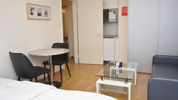 Habitación de hotel con una cama, una mesa redonda con sillas, una mesita de centro de vidrio, una kitchenette con microondas y un armario.