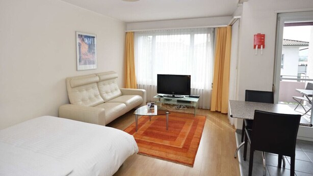 A modern hotel room with a bed, a sofa, a TV on a glass coffee table, a dining area with a table and chairs, and a large window with curtains.