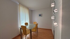 Een eetruimte met een glazen tafel en vier stoelen, een raam met zonwering, en een wandlamp.