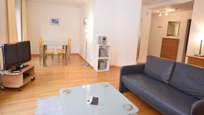 Een moderne woonkamer met een flatscreen-tv, een grijze bank, een eettafel met stoelen en een klein kastje.