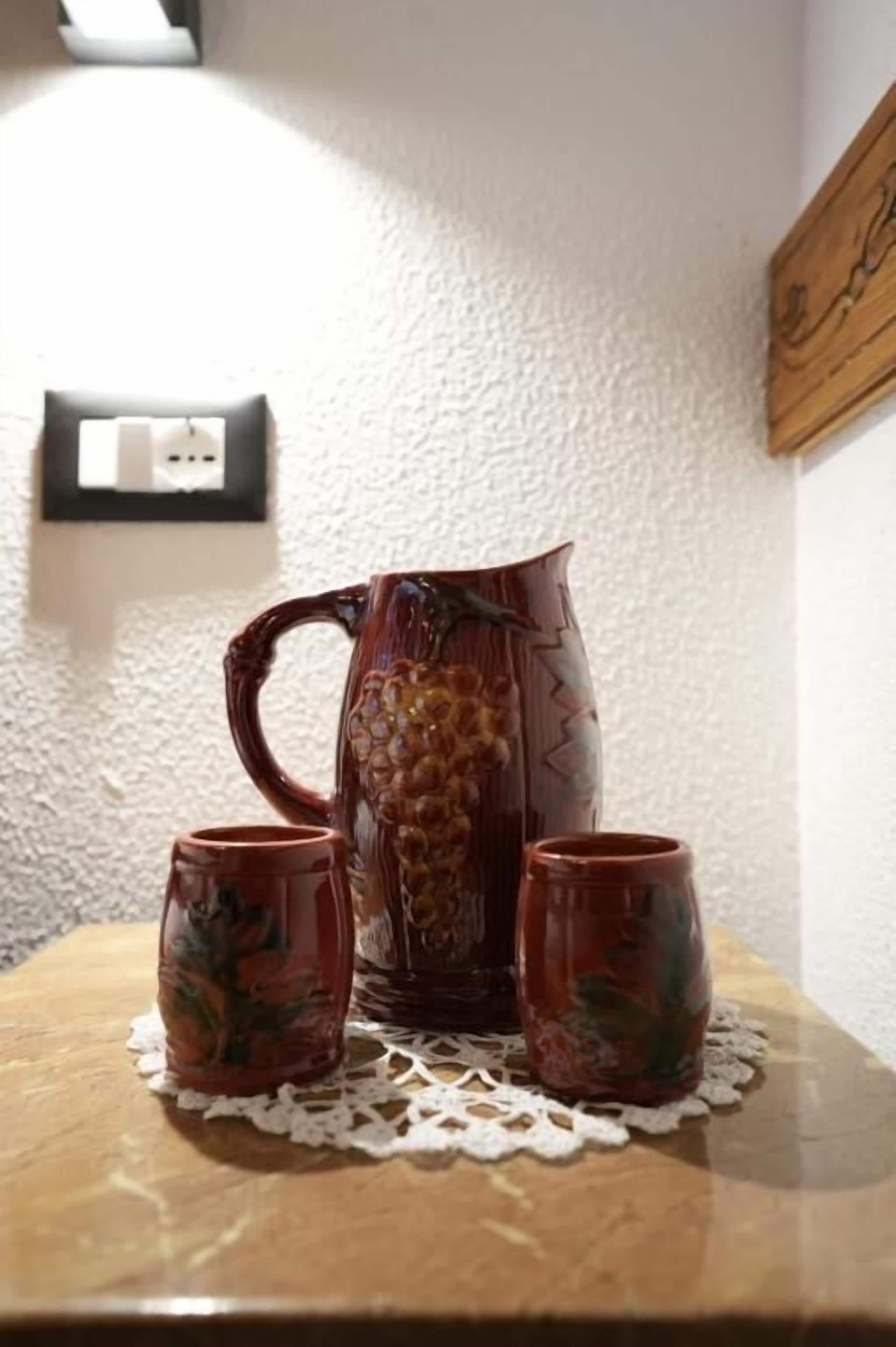 Un pichet en céramique et deux tasses assorties sont posés sur une table en bois, sur laquelle se trouve une nappe en dentelle blanche.