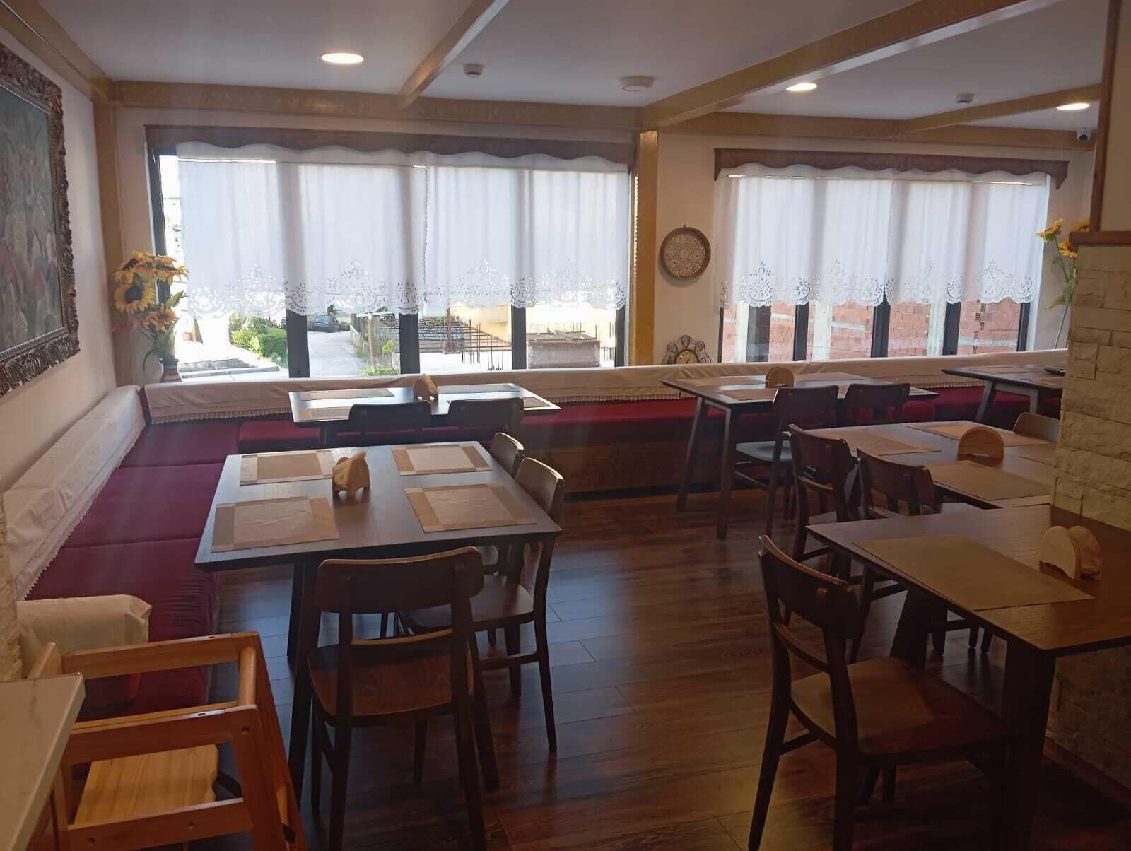 A restaurant with wooden tables and chairs, large windows with lace curtains, and a view of the street outside.