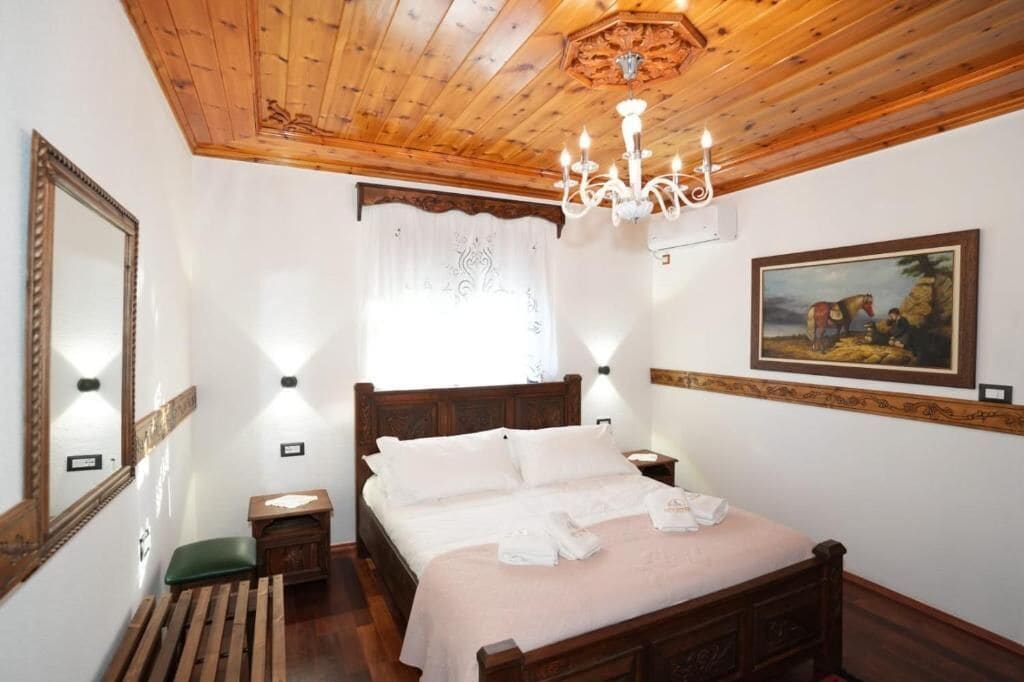 Un dormitorio con techo de madera, un candelabro, una cama con ropa de cama blanca, un banco verde, una mesita de noche de madera y un cuadro en la pared.