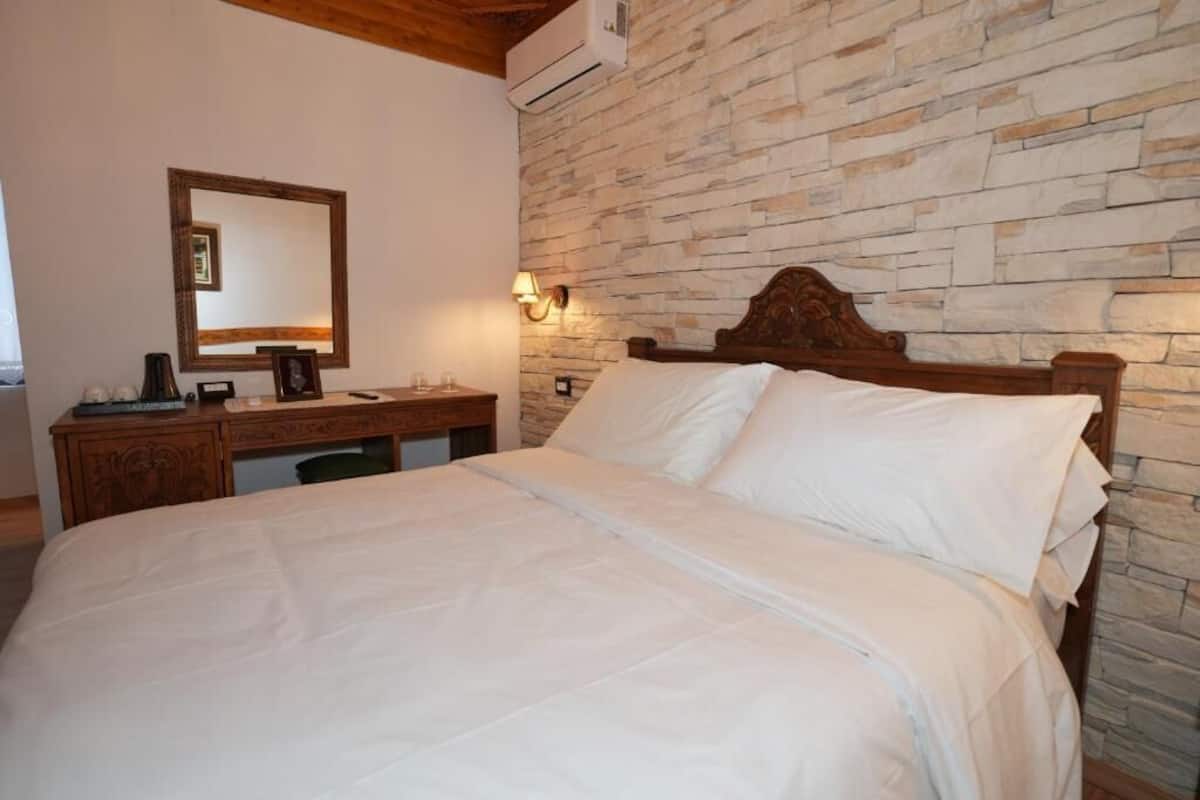 A bedroom with a stone wall, a wooden bed frame, a desk with a chair, and a mirror on the wall.