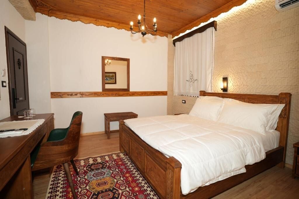 Een slaapkamer met een houten bed, een bureau met stoel, een klein tafeltje en een vloerkleed met dessin.
