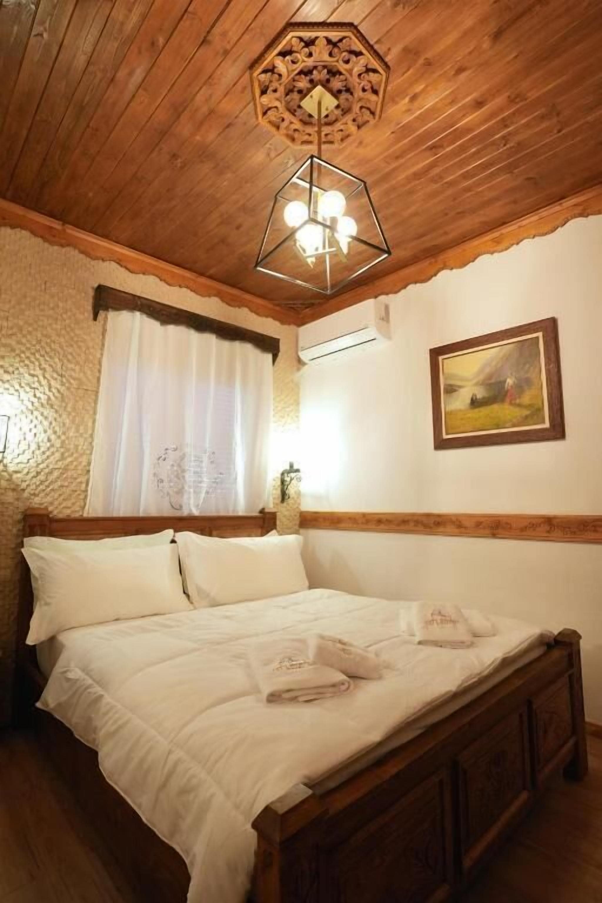 A wooden ceiling with a geometric light fixture, a bed with white linens, a window with white curtains, and a framed picture on the wall.