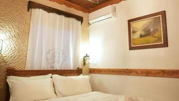 A wooden ceiling with a geometric light fixture, a bed with white linens, a window with white curtains, and a framed picture on the wall.