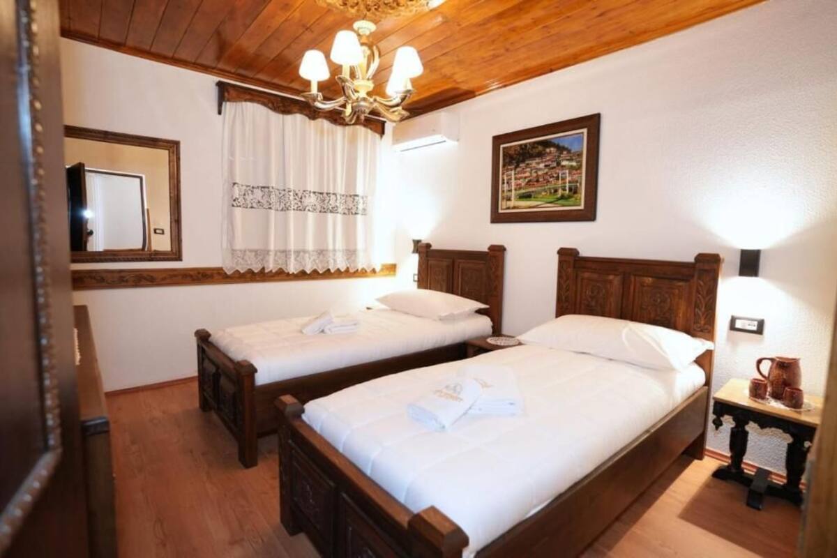 A room with two wooden beds, a chandelier, a mirror, a picture on the wall, and a small table with a pitcher and cups.