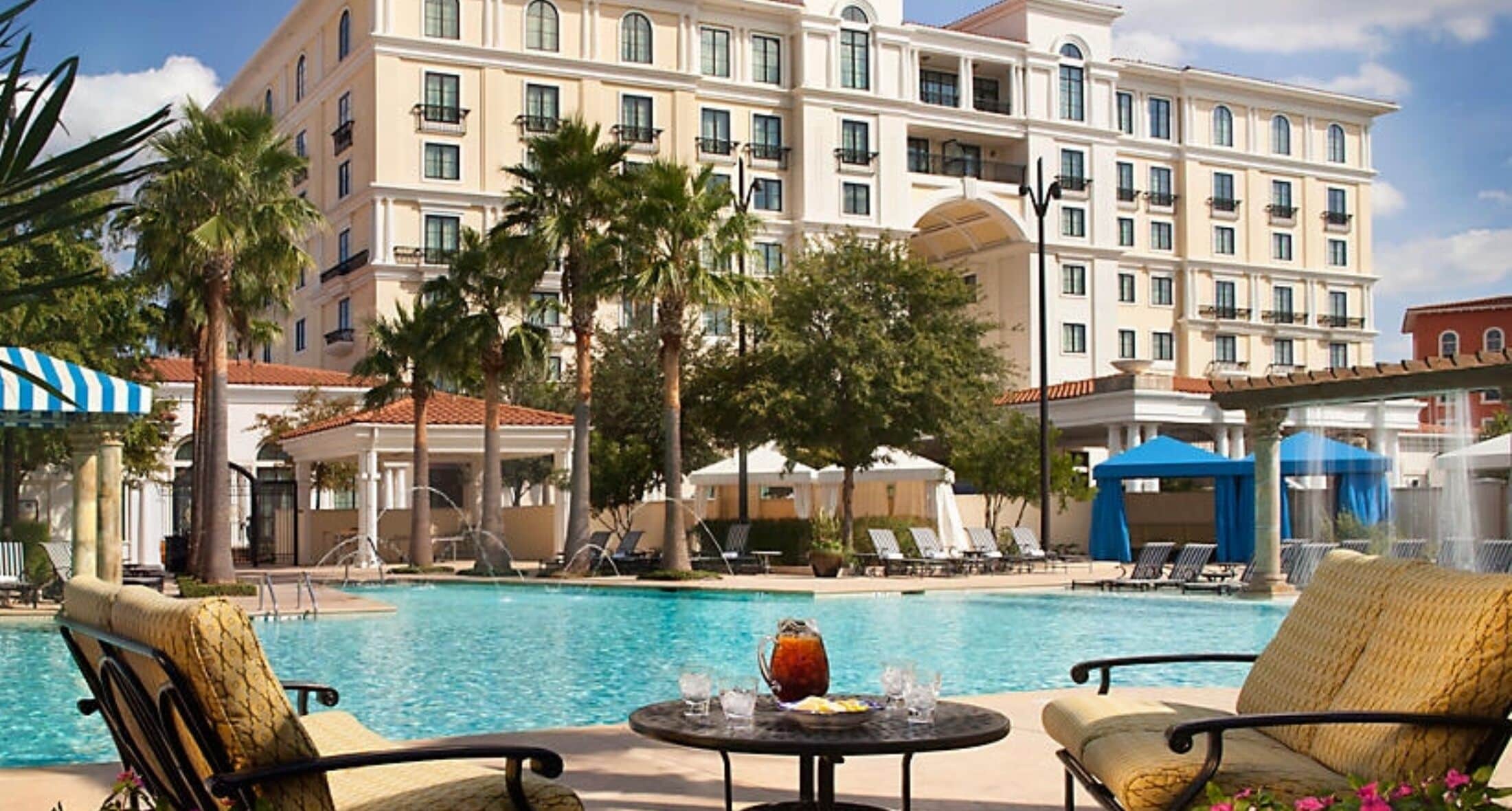 Zona de piscina de hotel con sillones reclinables, una mesa con bebidas y un edificio grande al fondo.