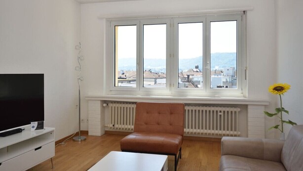 Una sala de estar con un ventanal grande, un televisor de pantalla plana, un sillón marrón, una mesa blanca y un sofá.
