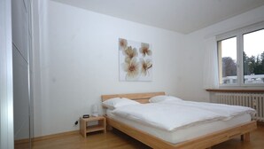 Un dormitorio con cama de madera, ropa de cama blanca, una mesita de noche con lámpara y una ventana con vistas a árboles.