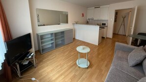 Een moderne woonkamer met een flatscreen televisie, een grijze bank, een klein rond tafeltje en een keuken met witte kasten en een aanrecht.
