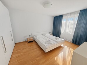 Quarto moderno com uma cama grande, soalho de madeira, mesa de cabeceira e janela com cortinas.