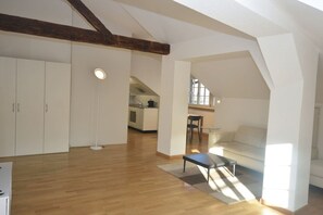 An open-plan living area with a wooden floor, a sofa, a coffee table, and a floor lamp. There is a kitchen in the background with a dining area. - Loft in the historic district (Symbol 4) (Zürich)