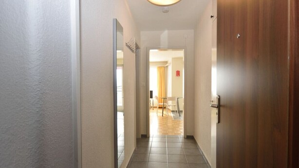 A hallway with a wooden door, a mirror, and a view into a dining area with a yellow curtain. - Very central & bright studio (Vineyard 51) (Zürich)