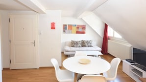 Et moderne, lyst værelse med en hvid dør, et hvidt bord med stole, en sofa med blomstrede puder og et fladskærms-tv på et hvidt stativ.
