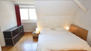 Et soveværelse med seng, sengeborde, en radiator, et vindue med gardiner og et opbevaringsmøbel.