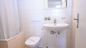 Et lille, rent badeværelse med hvide fliser, et hvidt toilet, en vask med et spejl over og et brusebadeforhæng.