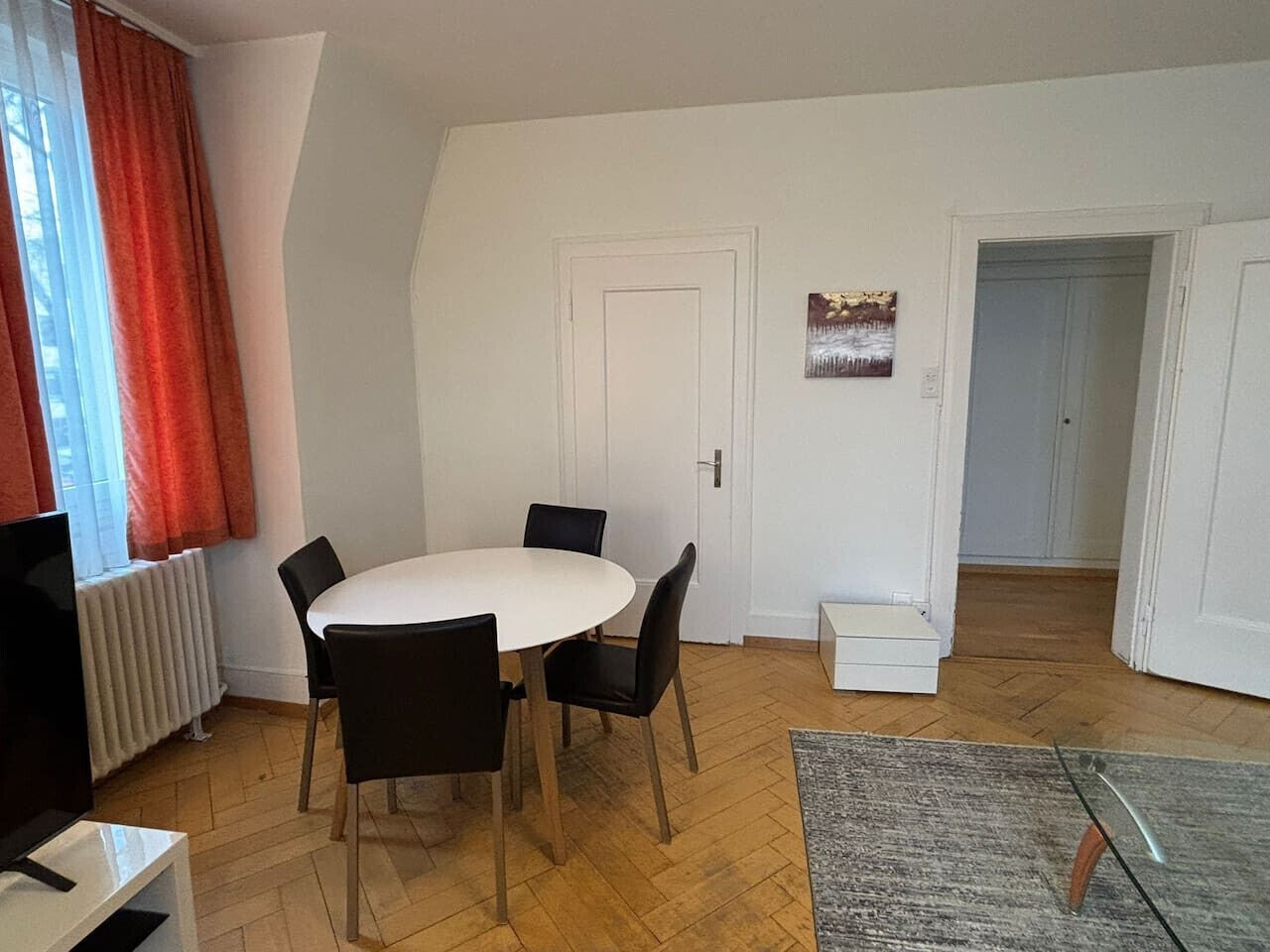 Un comedor con una mesa blanca y cuatro sillas, un radiador y una puerta con un escalón pequeño que lleva a otra habitación.